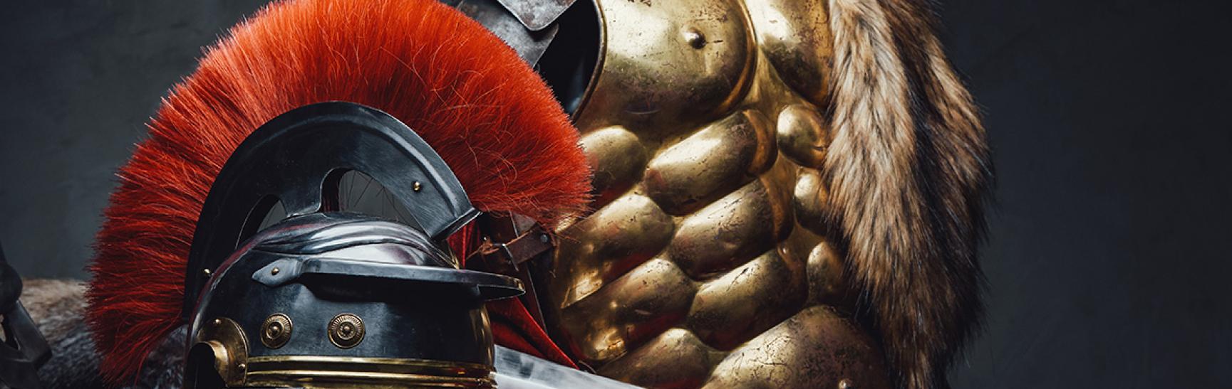 Image of Roman armour arranged casually as if just used: a metal breastplate and helmet with a red plume rest on top of a red cloak and some furs. There's also a gladius (shortsword), without any sheath or scabbard..