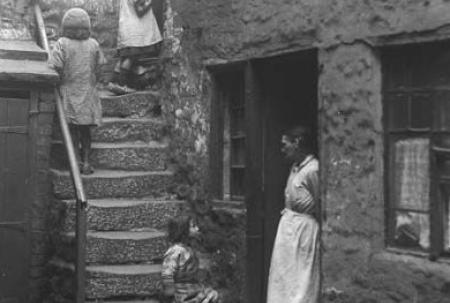 black and white photo of woman in white apron standing in doorway of small stone house. 3 children are sitting or standing on stone steps running up the wall next to the door.